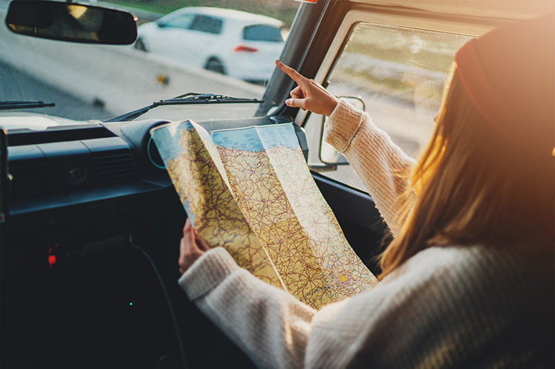 A person in a sweater and hat sits in a car passenger seat, holding a map while pointing out the windshield, suggesting they are navigating or giving directions during a road trip. Sunlight streams through the window.