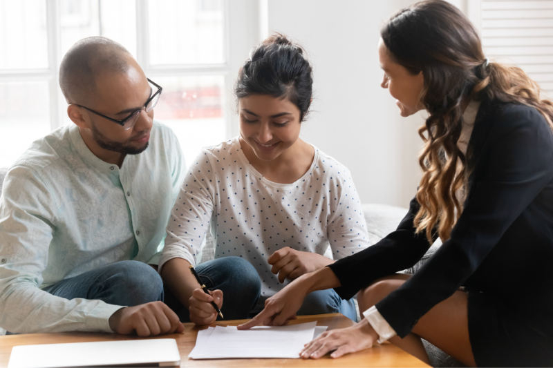 Home 1 A woman in business attire points to a document on a table while a seated man and woman listen and review the paper, appearing to discuss or sign paperwork together in a bright room.