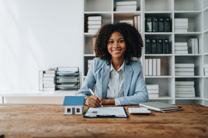 Home 6 A woman in a light blue blazer sits at a desk, smiling, with documents, a pen, a small house model, a notebook, and a calculator in front of her. Bookshelves with files and books are in the background.